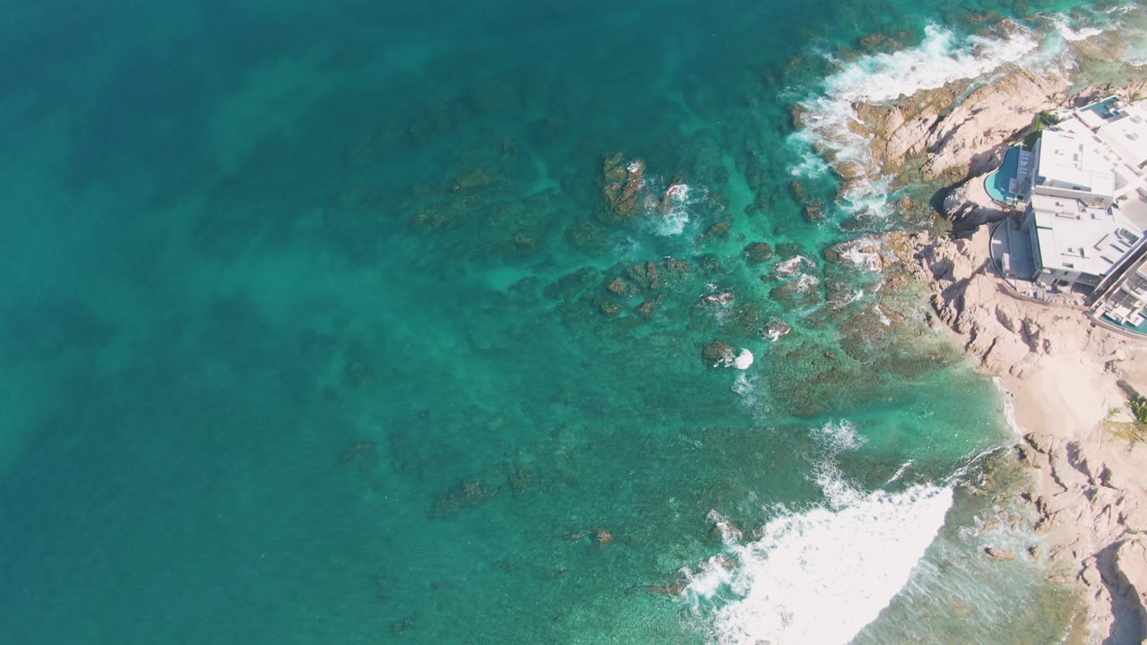 At one second, the El Tule Beach aerial view shows a fresh band of white foam spreading over rocks near the sand, with clear water around it and no visible people.