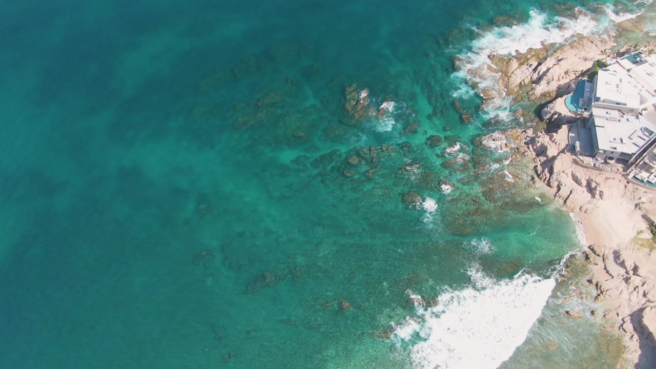At two seconds, the overhead El Tule Beach frame holds open water across most of the image while surf breaks near the rocky right edge and coastal buildings remain partly visible, with no visible people.