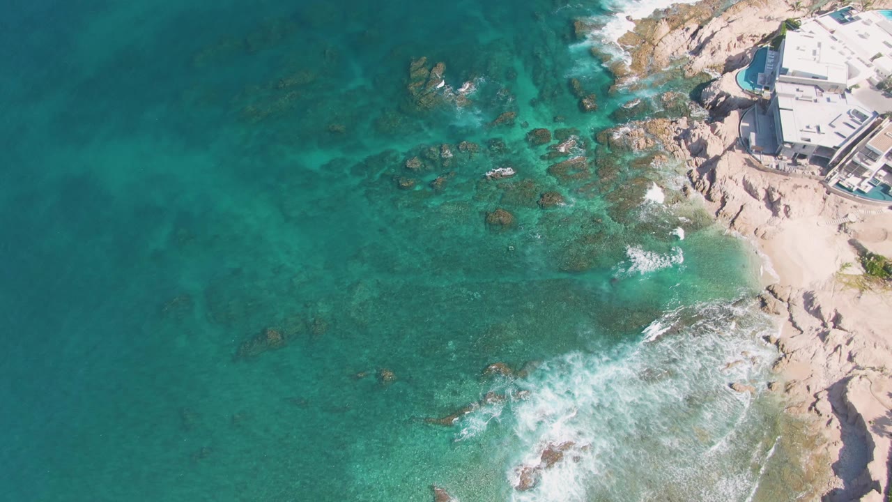 Aerial frame at El Tule Beach, Los Cabos showing clear green-blue water over rocks, a curved band of white surf lower right, pale stone shoreline, buildings at the top right, and no visible people.