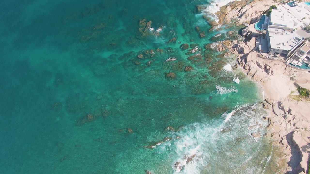 High overhead view of El Tule Beach in Los Cabos with darker underwater rock patches across the center, bright surf at the lower right, sandy rock ledges on the right, and no visible people.