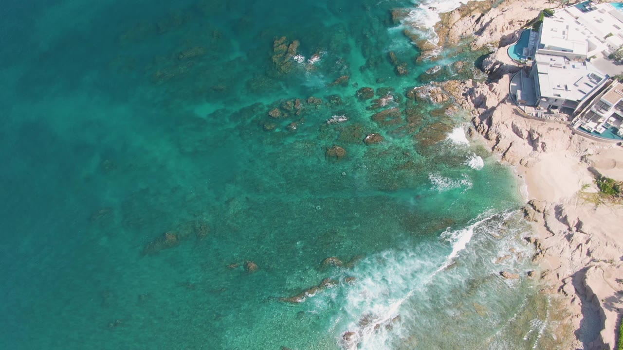 El Tule Beach aerial still with open turquoise water on the left, clustered rocks in the midground, white foam thinning near the lower right, coastal buildings at the upper right, and no visible people.