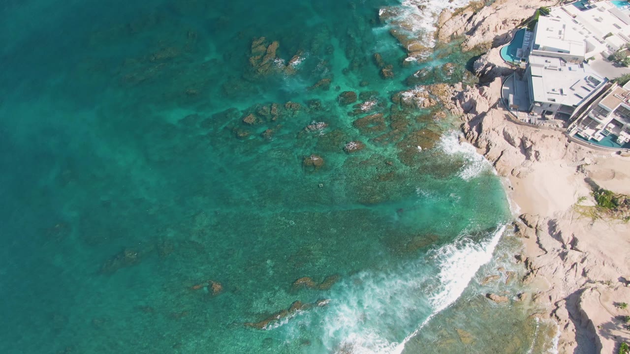 Aerial frame from El Tule Beach Los Cabos showing turquoise water over submerged rocks, white surf at the sand, and a light coastal building at the upper right with no visible people.