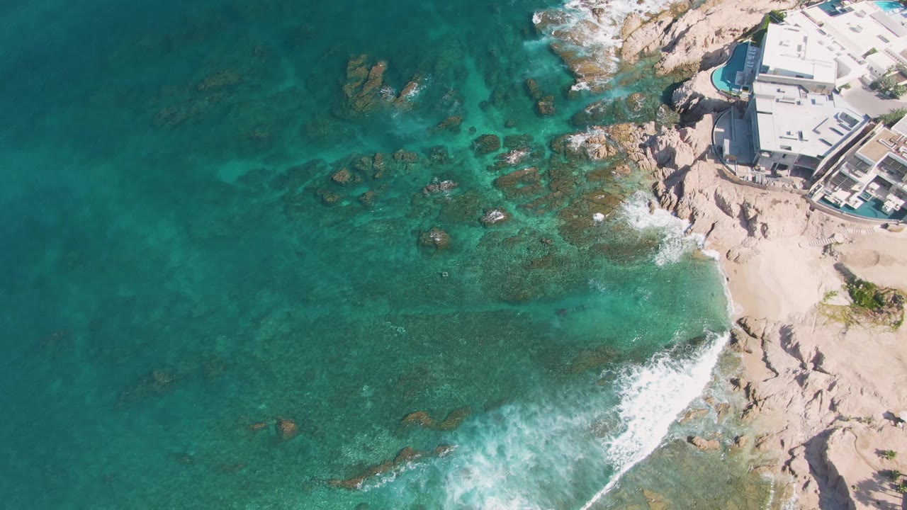 Aerial frame from El Tule Beach Los Cabos with foamy water curling toward pale sand, darker rocks visible under the turquoise surface, and the coastal building cropped at the upper right with no visible people.