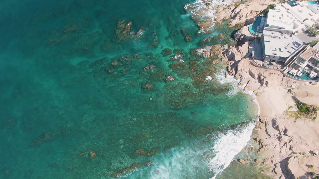 Aerial frame from El Tule Beach Los Cabos showing a broader patch of shallow turquoise water, surf breaking along the lower right beach, and the light-colored coastal building at the top right with no visible people.