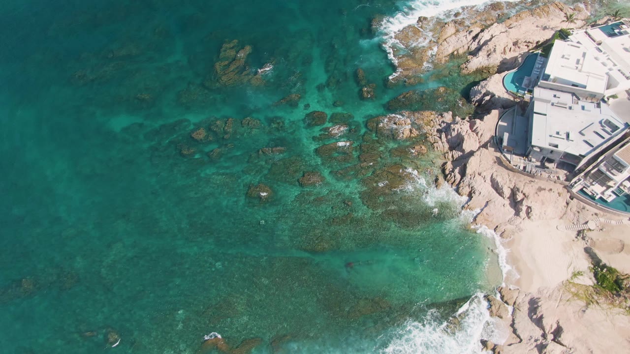 Aerial view over El Tule Beach, Los Cabos, with turquoise water covering dark rocks, white surf on pale shore, a small sandy area, a modern building at right, and no visible people.