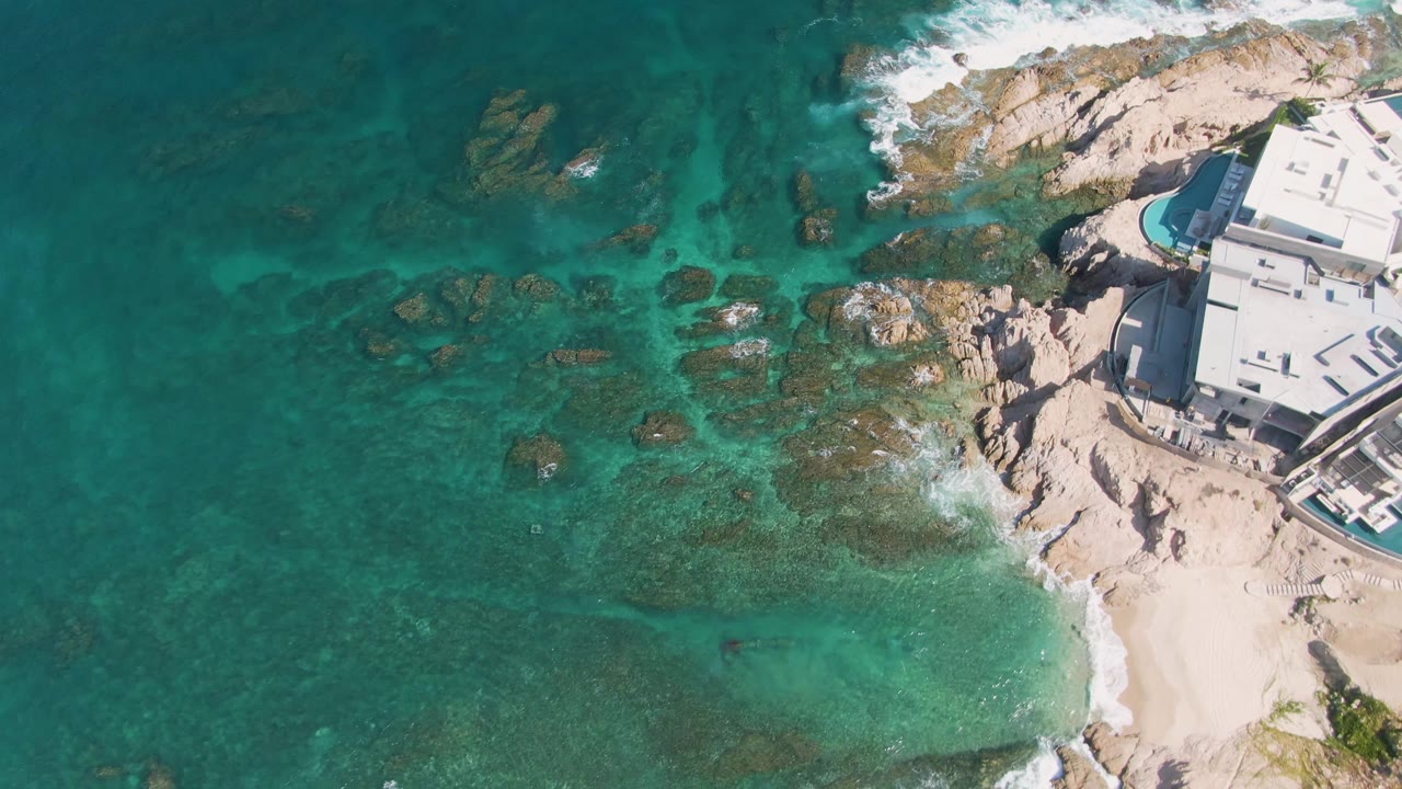 Overhead view at El Tule Beach, Los Cabos, with turquoise water darkening around rocks, a band of white foam on the upper shoreline, sand below, the building cropped at right, and no visible people.