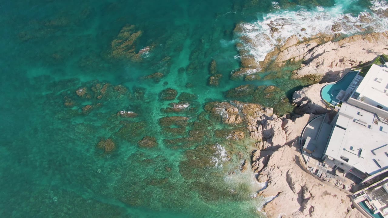 Aerial frame at El Tule Beach in Los Cabos showing more of the rocky shallows, white foam along the upper shoreline, the building on the right edge, and no visible people.