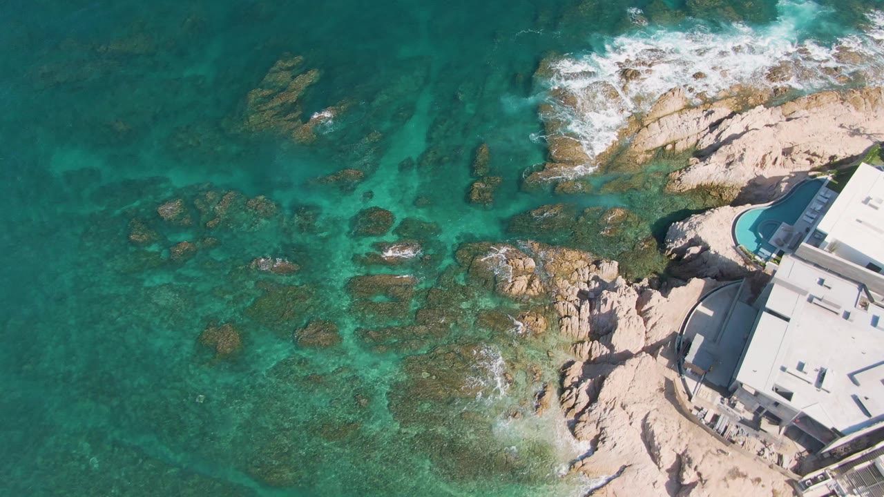 Aerial view over El Tule Beach in Los Cabos with surf spread across the upper rocks, transparent water in the center, the white building cropped at right, and no visible people.