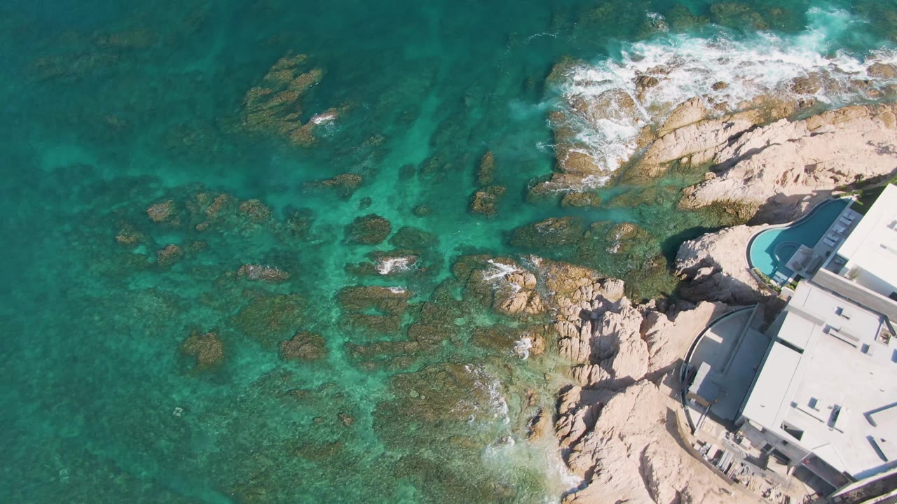 Mid-clip aerial frame at El Tule Beach in Los Cabos, where clear water reveals rocks below, foam collects near the shore, the curved pool area sits at right, and no visible people appear.