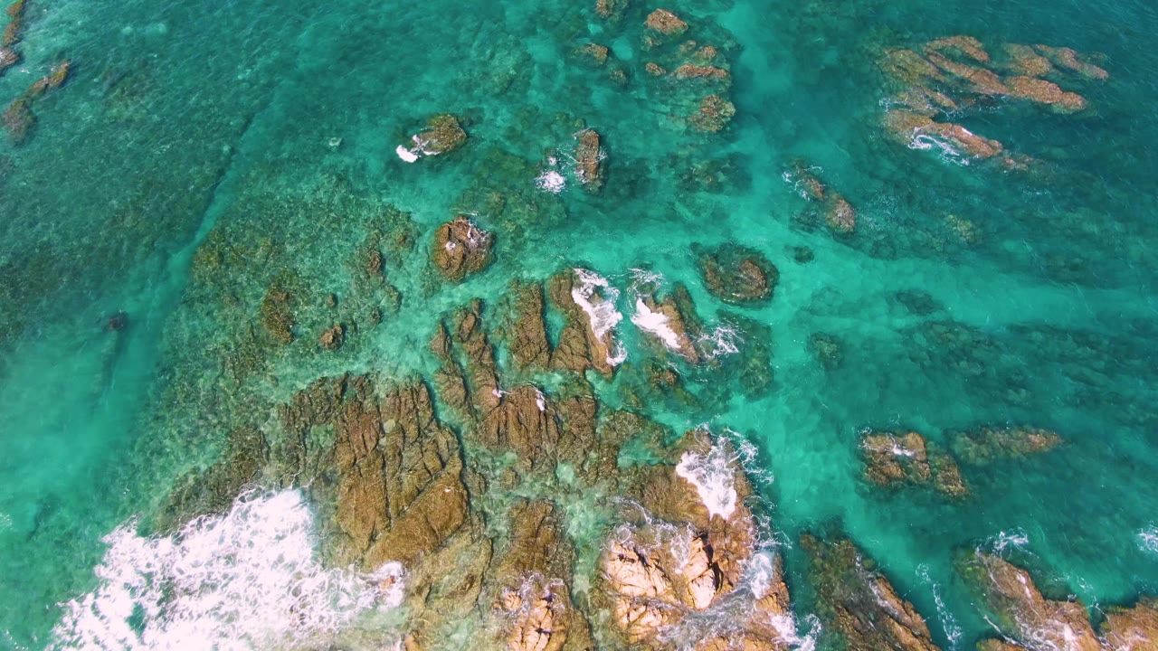 Aerial frame at El Tule Beach in Los Cabos showing clustered submerged rocks beneath turquoise water, with white surf wrapped around rocks near the lower center and no visible people.