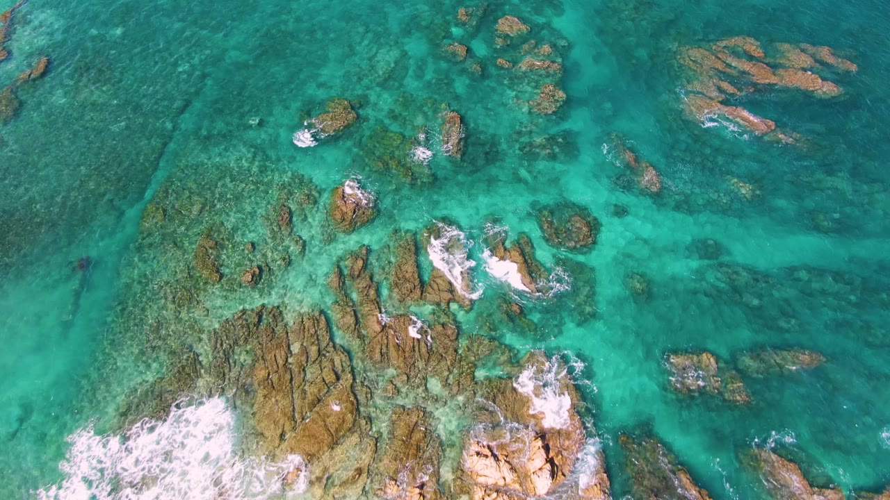 Top-down view of the El Tule Beach shoreline water in Los Cabos, where scattered rocks sit under clear turquoise shallows and foam breaks across the lower middle, with no visible people.