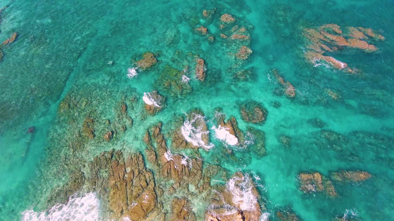 Overhead frame from El Tule Beach in Los Cabos with a rocky seabed visible through teal water and a larger band of white foam pushing in from the bottom edge, with no visible people.
