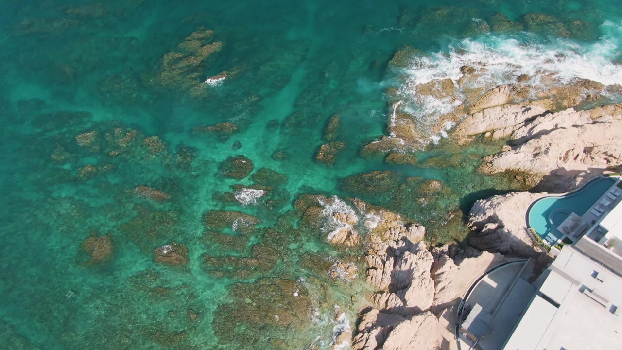 Overhead view at El Tule Beach in Los Cabos with clear turquoise water, submerged rocks, white surf on the upper rocks, and a curved pool building edge at right, with no visible people.