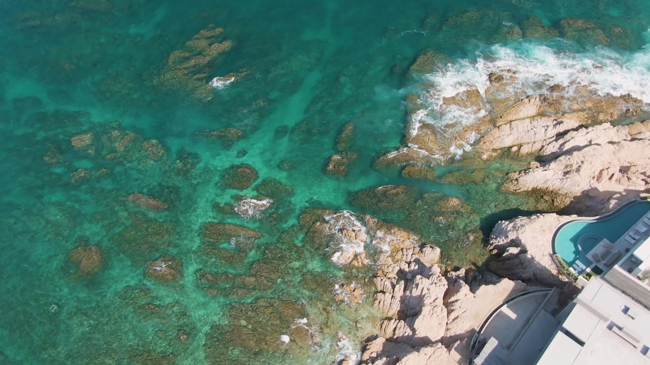 Aerial frame at El Tule Beach in Los Cabos showing shallow teal water over scattered rocks, wave foam breaking near the top, and the pool corner and building edge on the right, with no visible people.