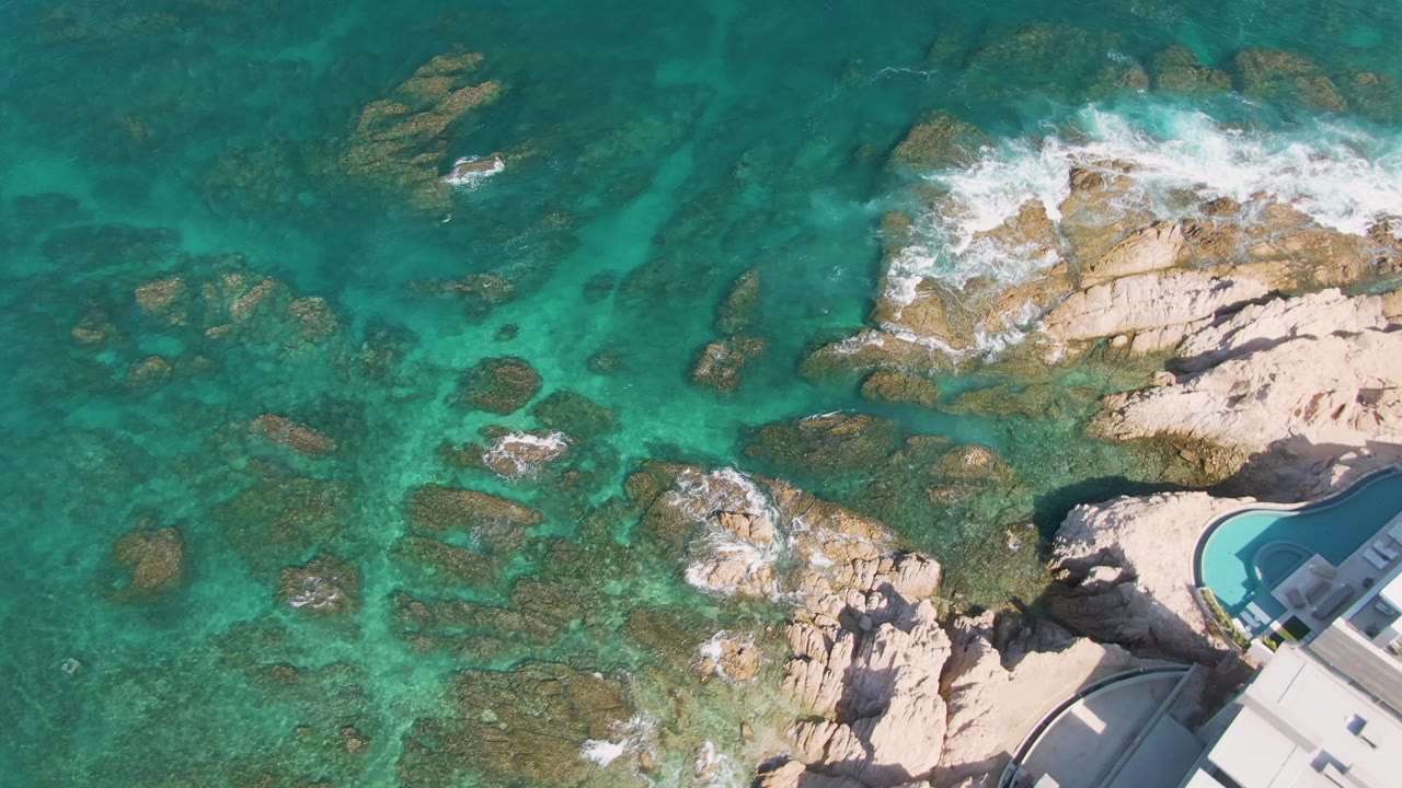 Overhead Los Cabos shoreline frame with turquoise ocean around tan rocks, fresh white foam at the upper right, and a light roof and curved pool edge entering from the right, with no visible people.