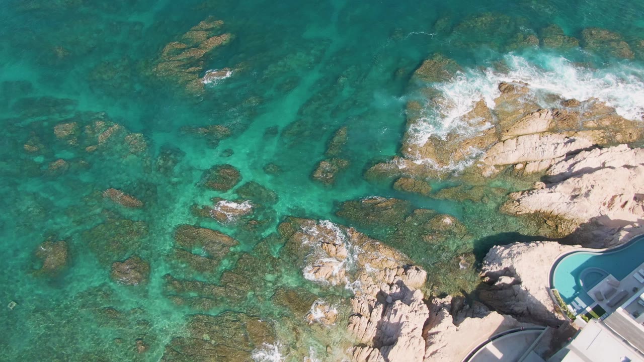 Aerial view at El Tule Beach in Los Cabos with more open turquoise water on the left, pale rocks below center, surf breaking across the upper rock shelf, and a partial pool building at right, with no visible people.