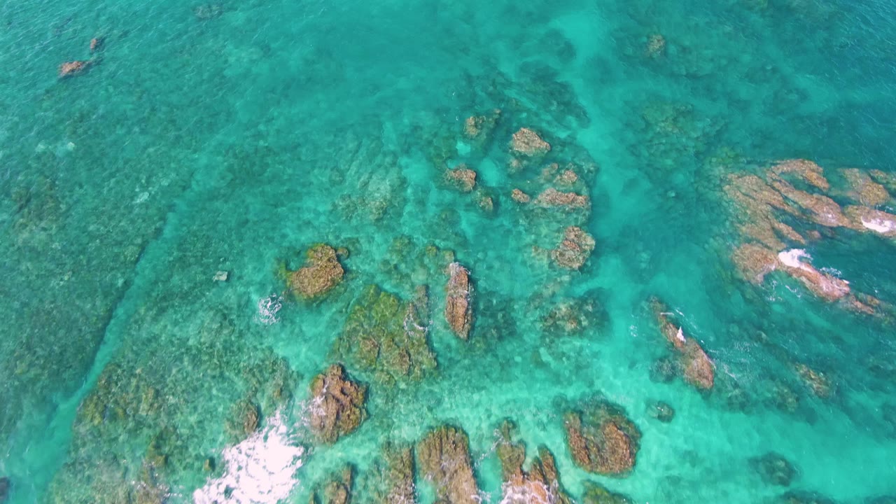 Aerial downward frame of clear turquoise water at El Tule Beach in Los Cabos, with tan rock clusters near the center and darker reef patches visible below the surface, no visible people.