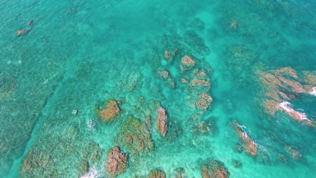 Clear turquoise ocean water at El Tule Beach in Los Cabos, with a broad reef patch on the left, scattered rock formations, and small whitecaps breaking on rocks to the right, no visible people.
