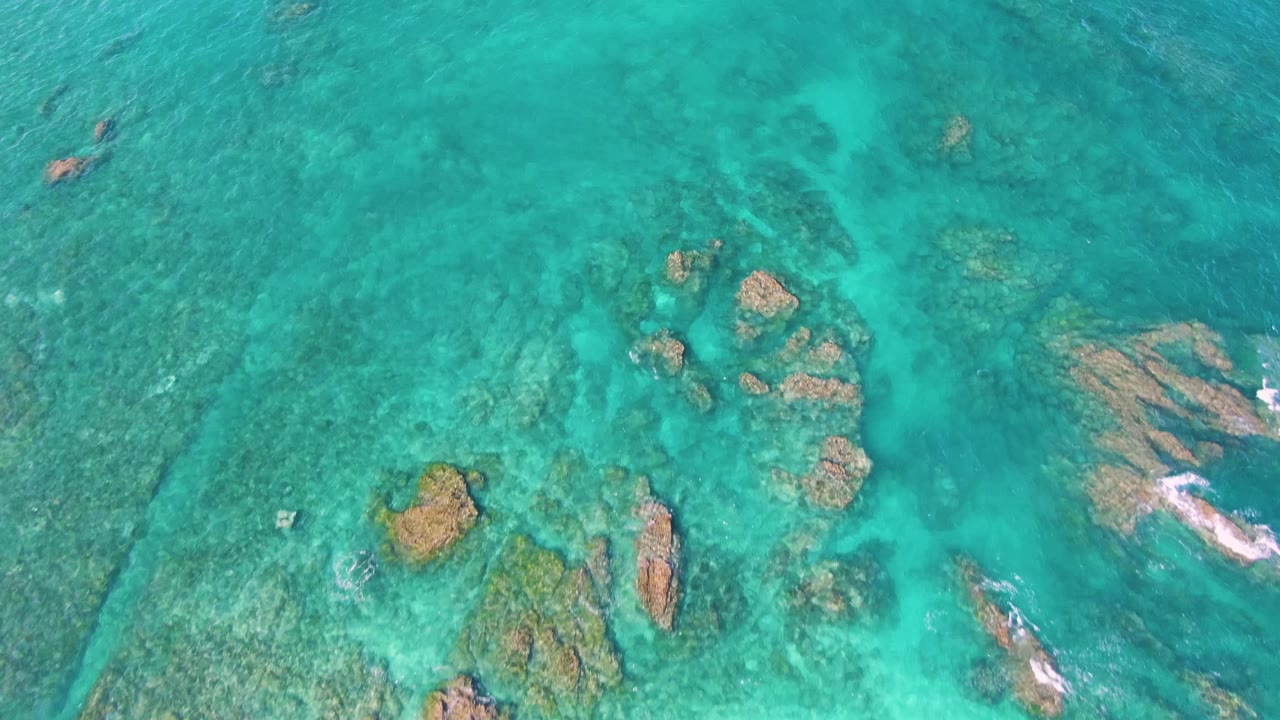 Top-down El Tule Beach water detail with turquoise shallows, scattered brown rock heads, darker underwater reef areas, and fresh white foam near the lower left edge, no visible people.