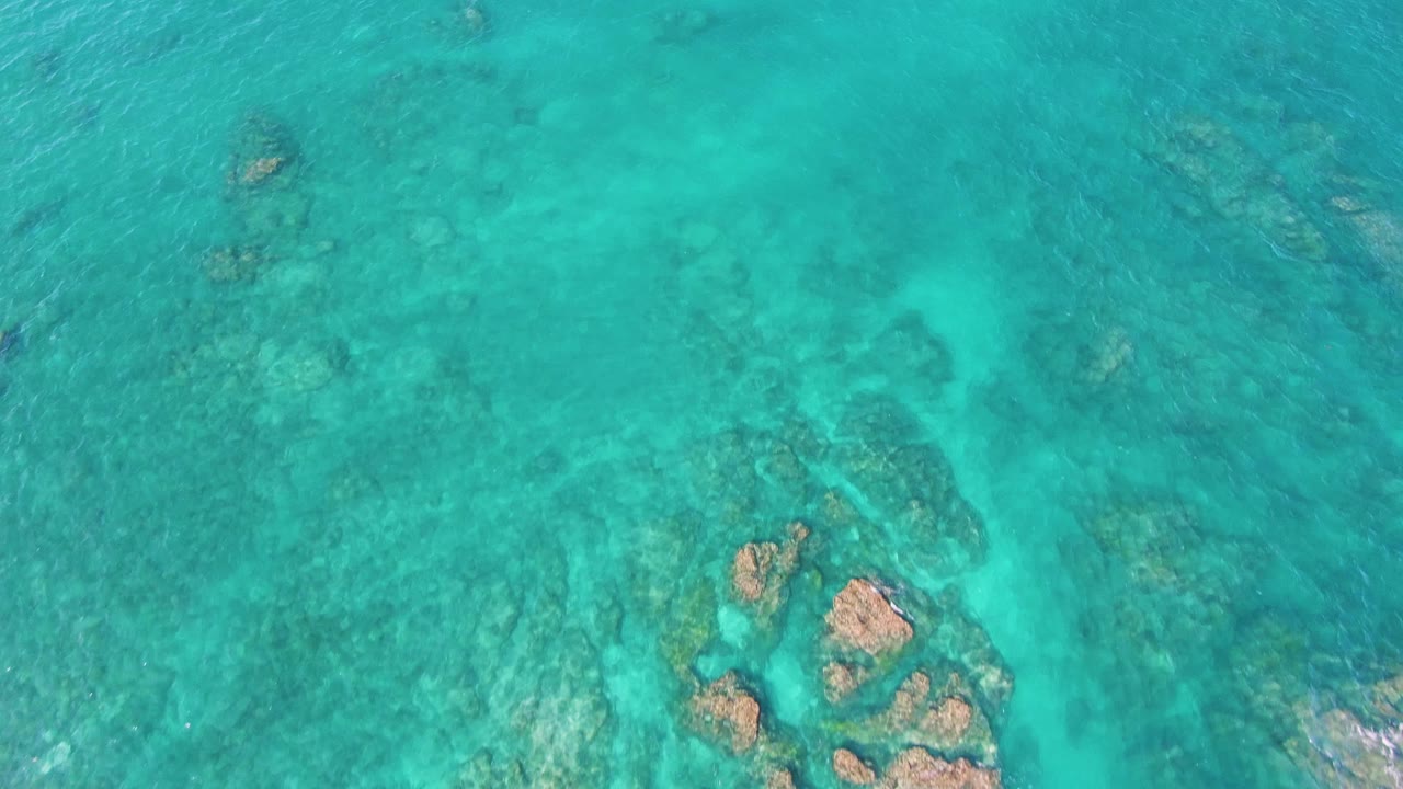 Clear turquoise water reveals a cluster of submerged brown rocks near the lower edge at El Tule Beach, Los Cabos, with no visible people.