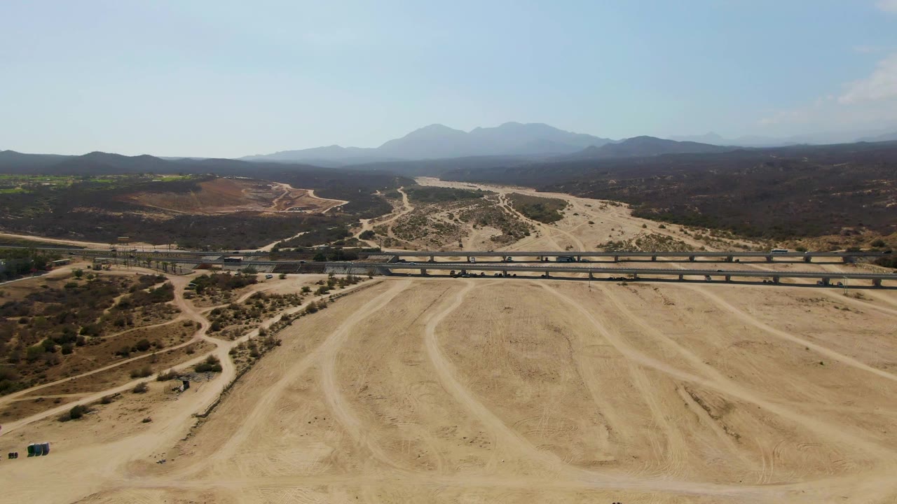 Aerial frame at El Tule Beach Los Cabos with the train farther along the rail bridge, broad sandy tire-marked ground below, dark dry hills in back, distant mountains, and no visible people.