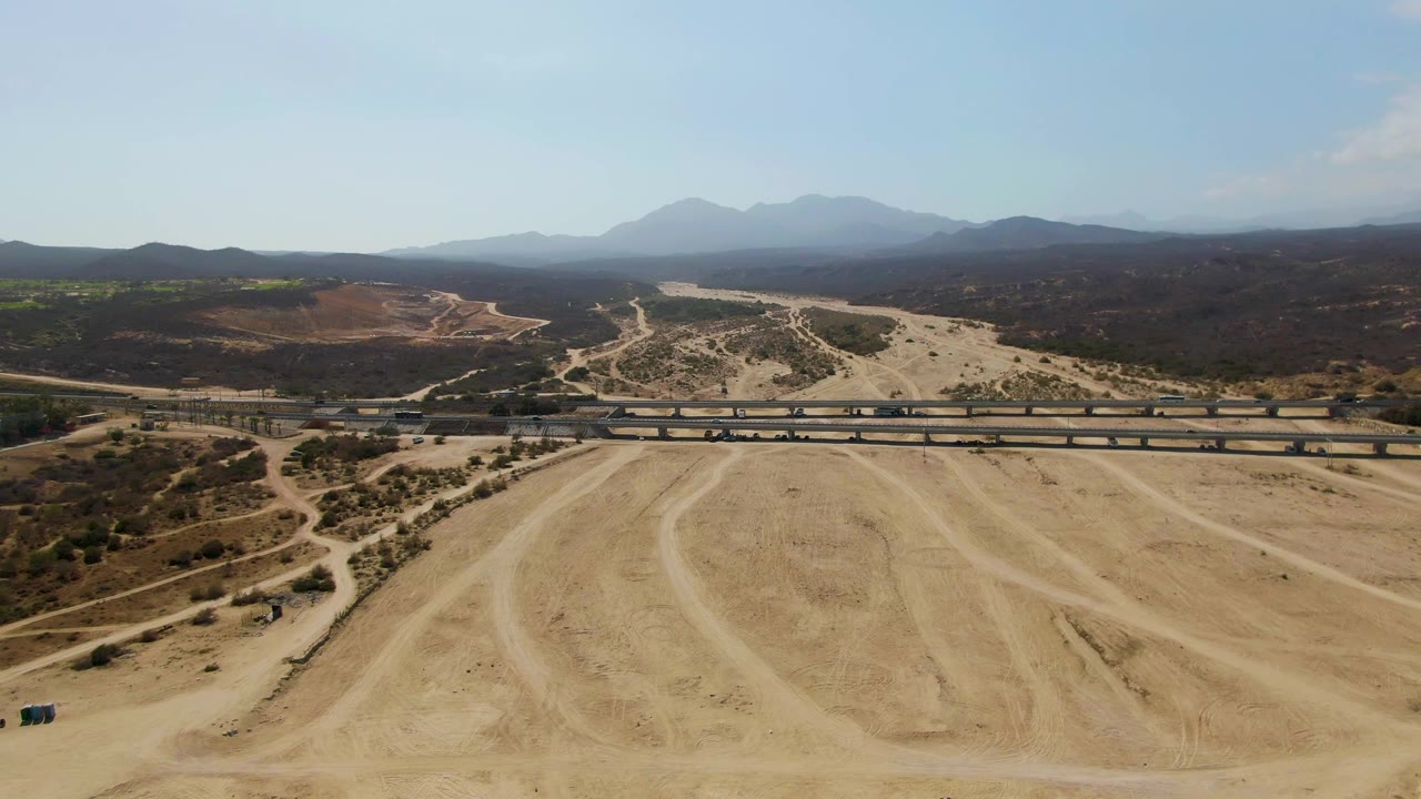 Wide aerial frame at El Tule Beach Los Cabos showing the train near the center-right section of the bridge, pale sand and curved tire marks in front, scrubby hills beyond, distant mountains, and no visible people.
