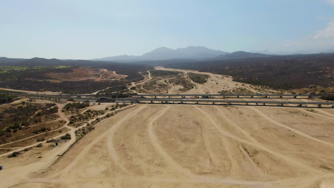 Aerial frame at El Tule Beach Los Cabos with the train advancing along the bridge, a large sandy quarry pad with track marks below, dark arid hills behind, hazy mountain silhouettes, and no visible people.