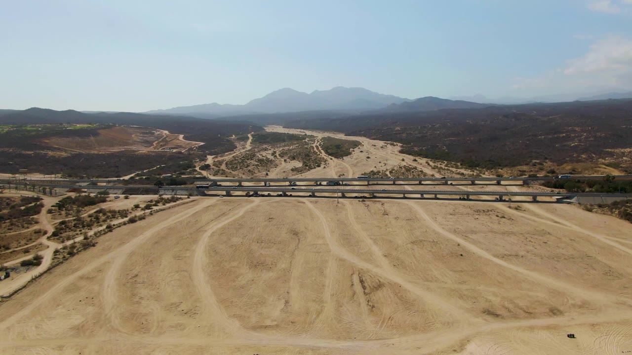 Aerial frame over El Tule Beach Los Cabos terrain with pale dirt tracks in the foreground, a bridge across the midground, hazy mountains beyond, and no visible people.