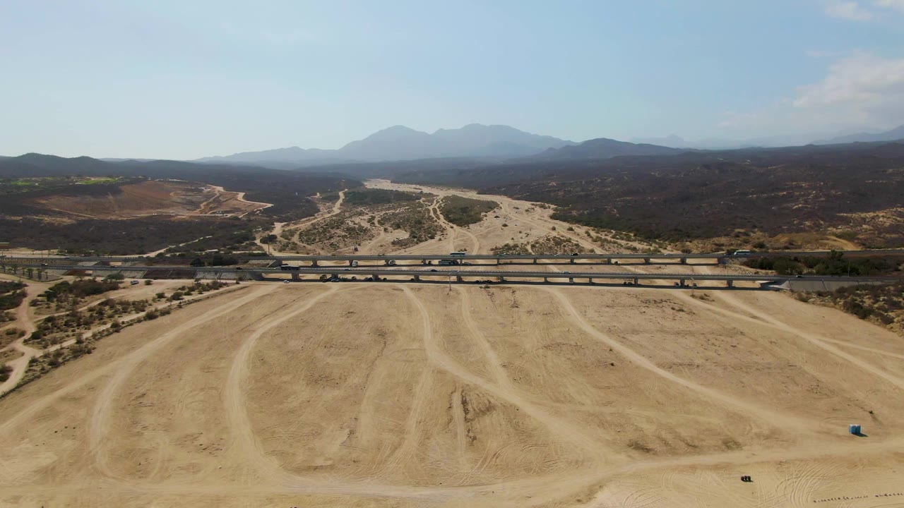 Wide drone view of circular dirt tracks with a road bridge and mountains beyond.