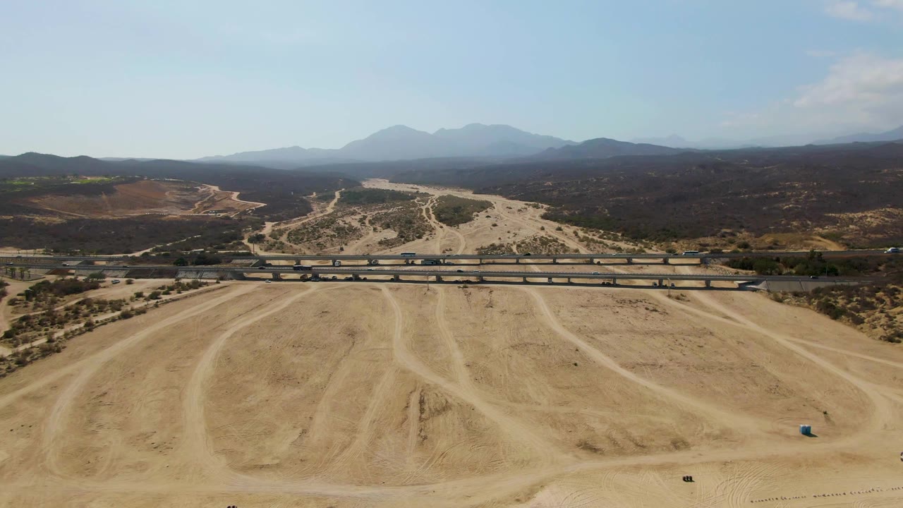Aerial frame from El Tule Beach Los Cabos with dry rutted ground in front, a horizontal road bridge across the scene, layered hills in the distance, and no visible people.