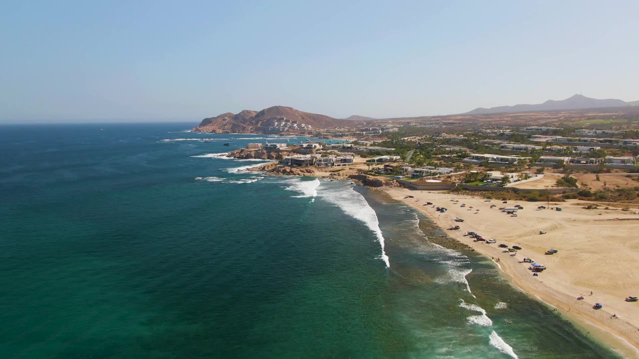 Aerial view over El Tule Beach in Los Cabos, with turquoise water on the left, white surf entering the sandy shore, parked cars on the beach, distant hills, and no visible people.