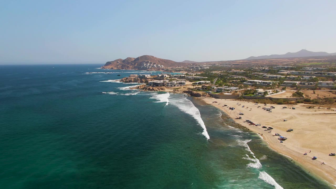 Drone view over El Tule Beach in Los Cabos, showing a bright white wave line angled toward the sand, deep blue-green water offshore, beach parking, low coastal development, distant hills, and no visible people.