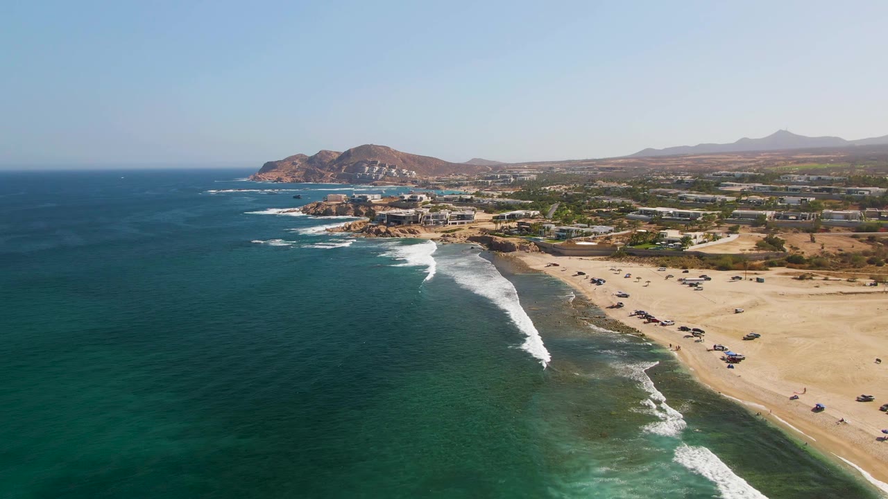 Aerial coastline frame at El Tule Beach in Los Cabos, with the surf line slightly farther down the beach, a broad band of sand with parked cars, low structures near the shore, hazy hills, and no visible people.