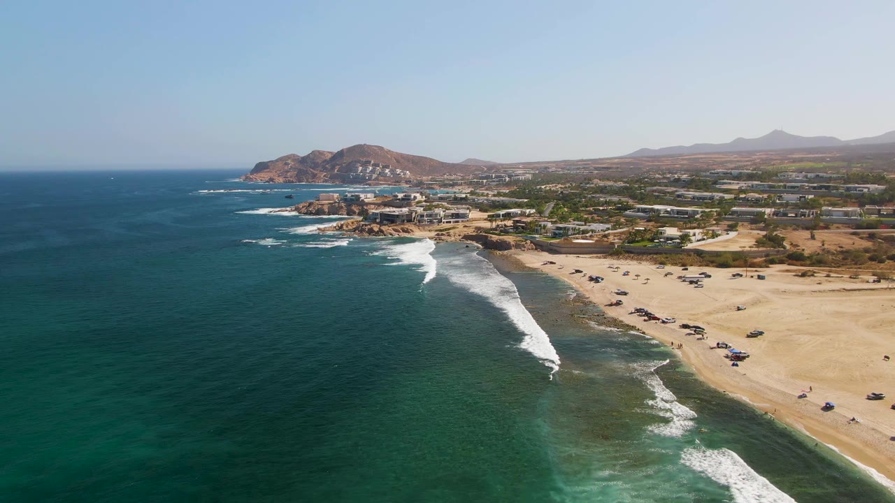 High aerial shot of surf washing toward a sandy beach and coastal hills.