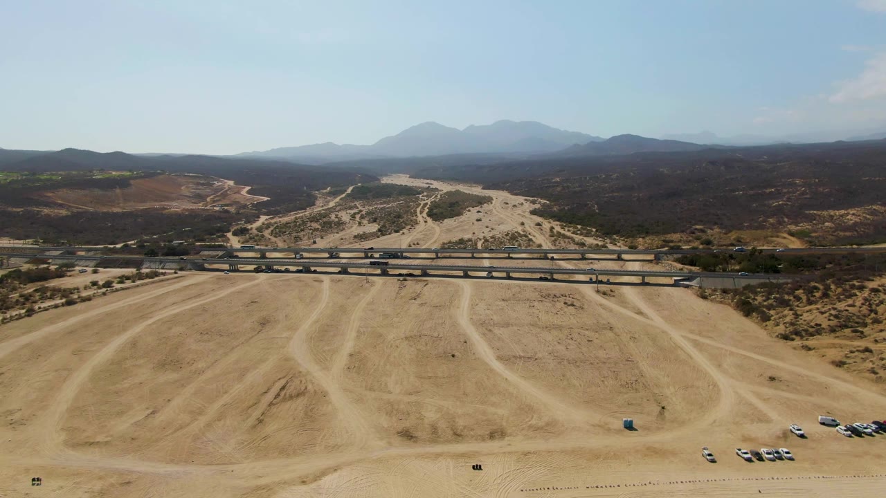 Aerial view over El Tule Beach Los Cabos showing a sandy parking area with tire tracks, a road overpass with vehicles, scrubland, distant mountains, and no visible people.