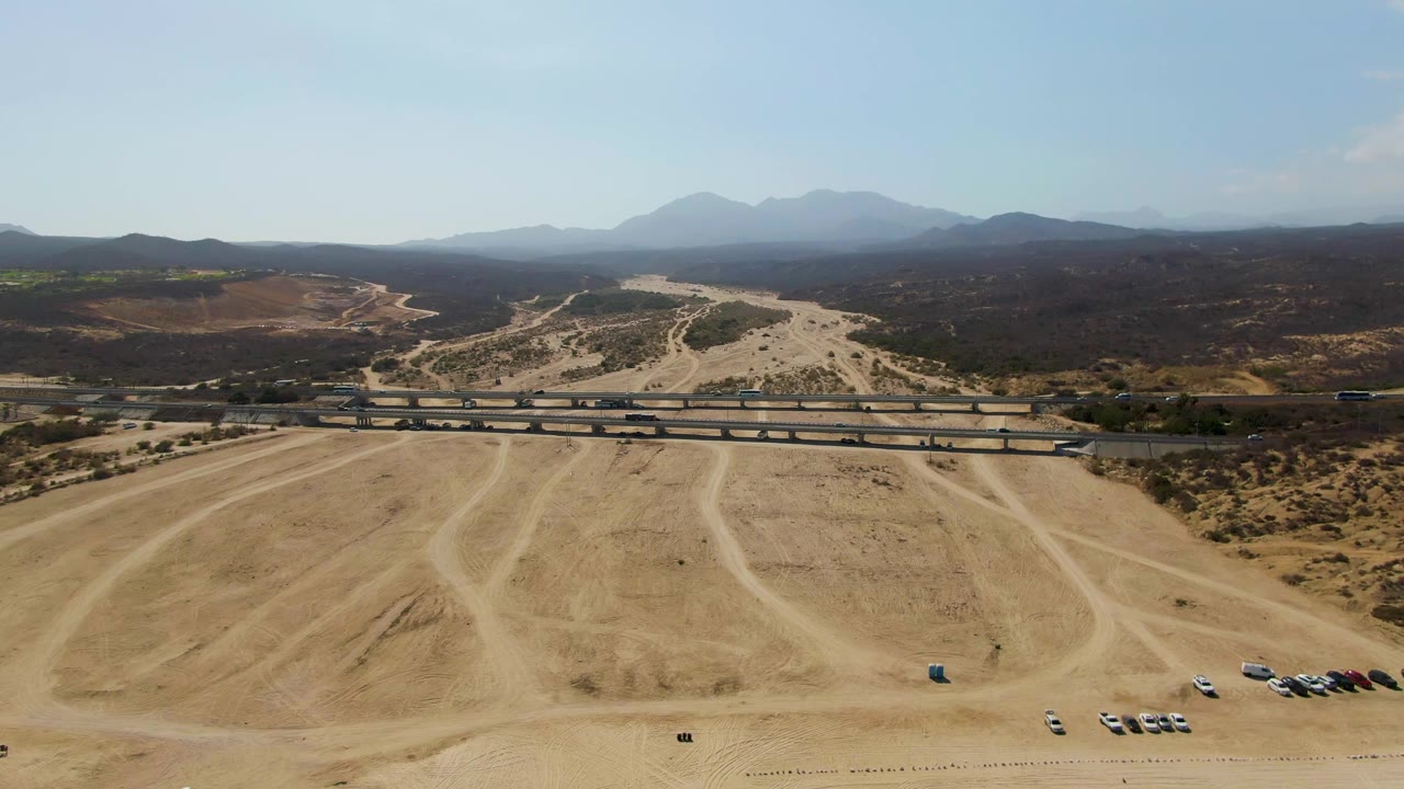 Wide drone view of dry dirt lot beside an overpass with vehicles and mountains beyond.