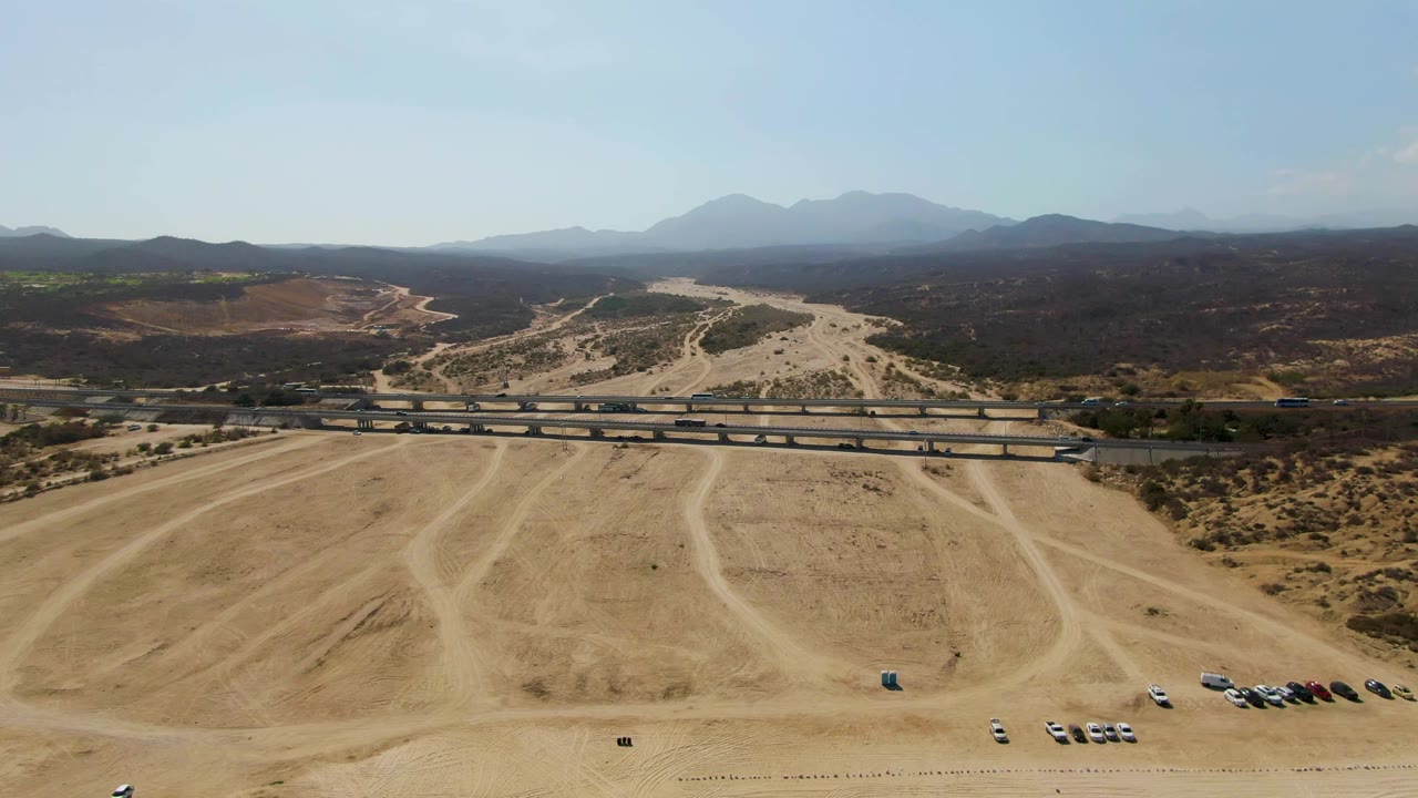 High aerial frame from El Tule Beach Los Cabos showing tire-marked sand in the foreground, small parked cars, overpass traffic, hazy mountain background, and no visible people.