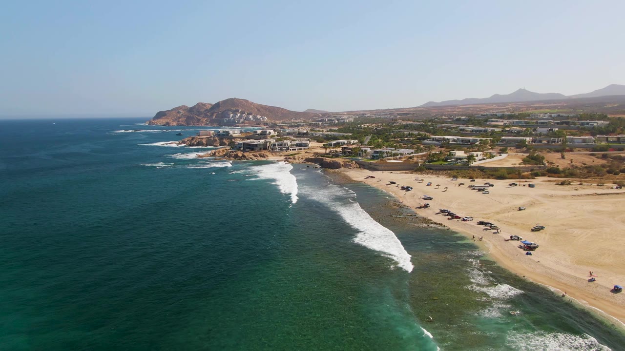 Aerial frame over El Tule Beach in Los Cabos showing deep blue ocean, turquoise shallows, white surf, sandy beach vehicles, coastal buildings, hills, and tiny visible people near the shoreline.