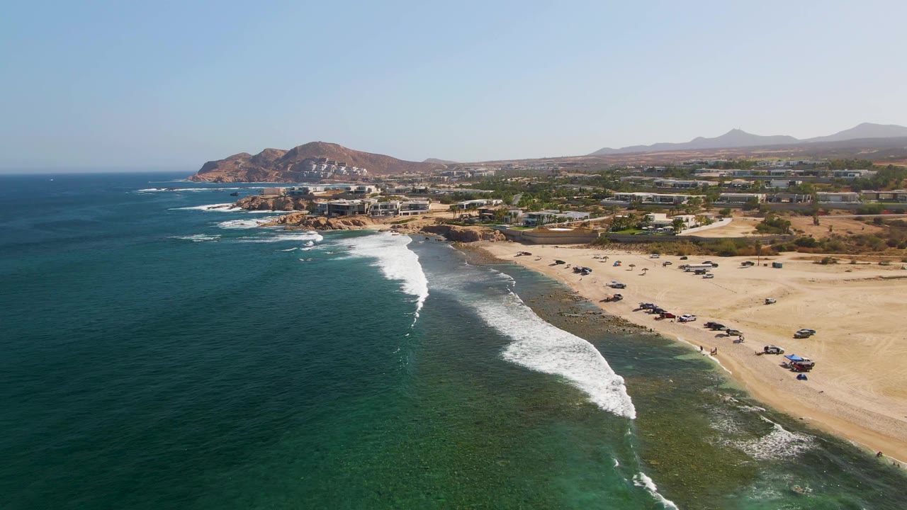 Wide aerial over El Tule Beach in Los Cabos with a clean horizon, darker open water at left, foamy surf near center, vehicle tracks and parked vehicles on the sand, and development backed by dry hills.