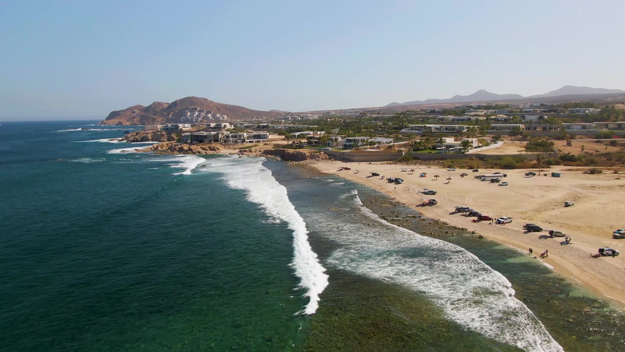 Aerial frame over El Tule Beach in Los Cabos, Mexico, with blue-green ocean at left, white surf reaching the sand, parked vehicles along the beach, low buildings beyond, hills in the distance, and no visible people.