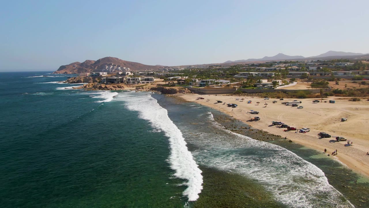 Drone-height view at El Tule Beach in Los Cabos, Mexico, where a broad white wave line pushes toward the lower edge, with beach vehicles to the right, coastal buildings behind them, distant hills, and no visible people.
