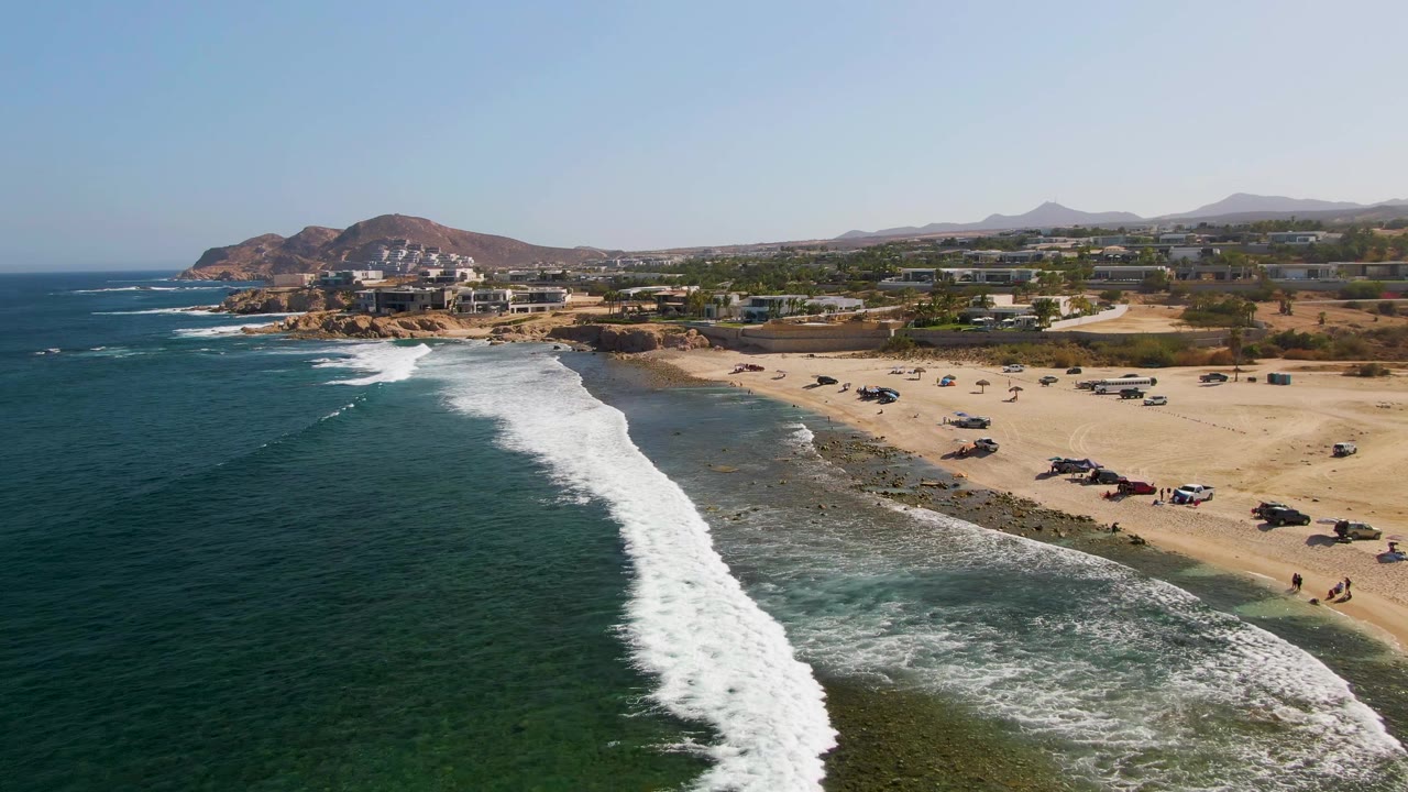 Aerial view of El Tule Beach in Los Cabos, Mexico, with the foamy breaker centered more prominently, dark ocean filling the left side, sandy parking areas and low structures on the right, hazy hills behind, and no visible people.