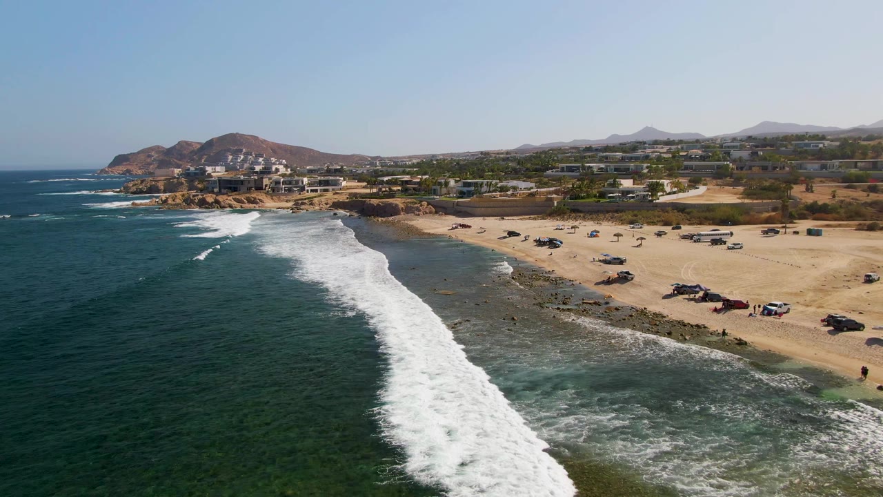 Wide aerial of surf rolling onto a beach with vehicles and coastal buildings