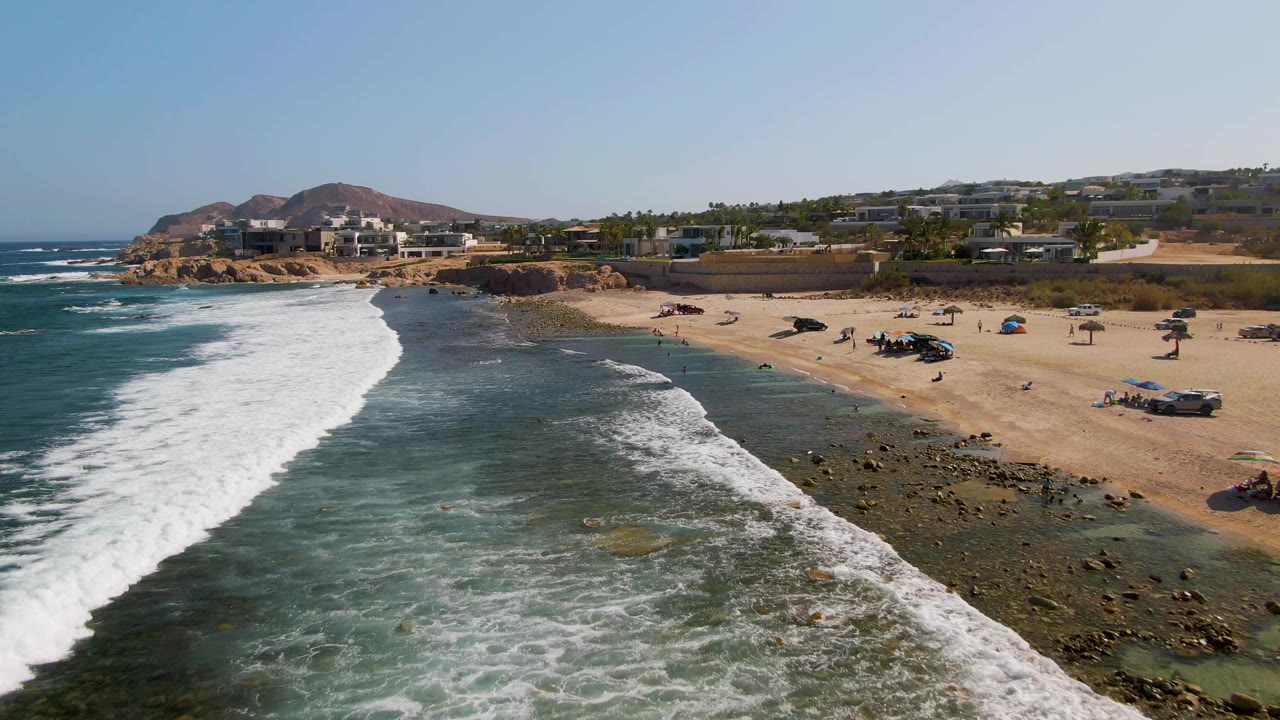 Aerial view at El Tule Beach Los Cabos showing white surf entering from the left, shallow rocky water, sandy beach, distant people, parked vehicles, coastal buildings, hills, and clear sky.