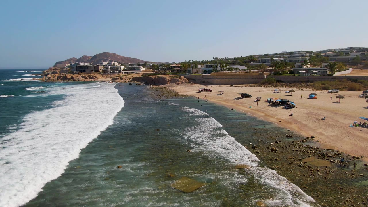 Drone view of a wave line crossing rocky shallows toward a sandy beach.