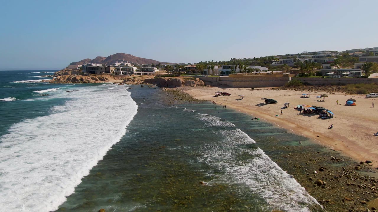 Aerial shoreline view at El Tule Beach Los Cabos where the surf has spread farther into white foam along the left side, with exposed rocks, sandy beach, distant people, vehicles, beach shade, hillside buildings, and blue sky.