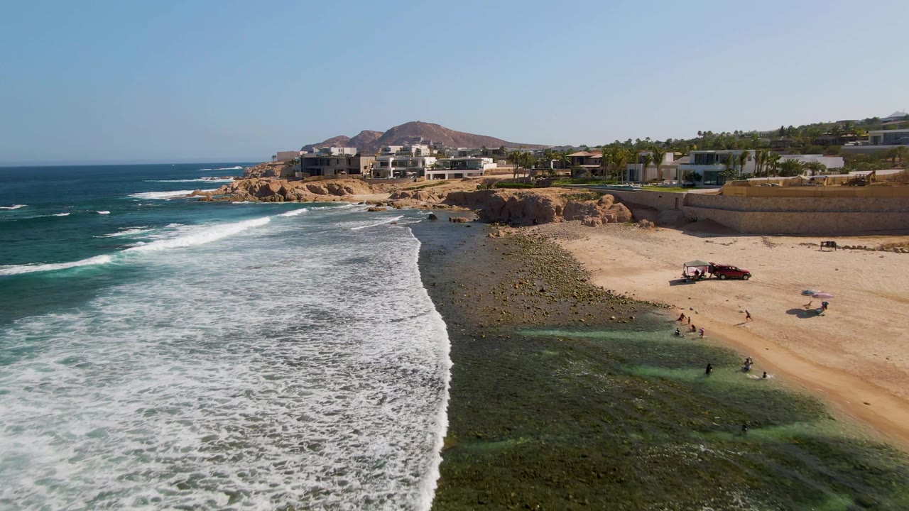 Aerial frame over El Tule Beach in Los Cabos showing white surf entering from the left, a sandy and rocky shoreline, small people in the shallows, beachfront buildings, cliffs, and a low hill under clear sky.