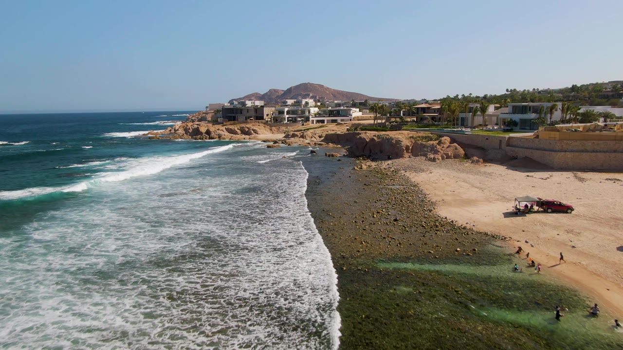 Waves break beside a rocky beach with people near the water and buildings on the bluff.