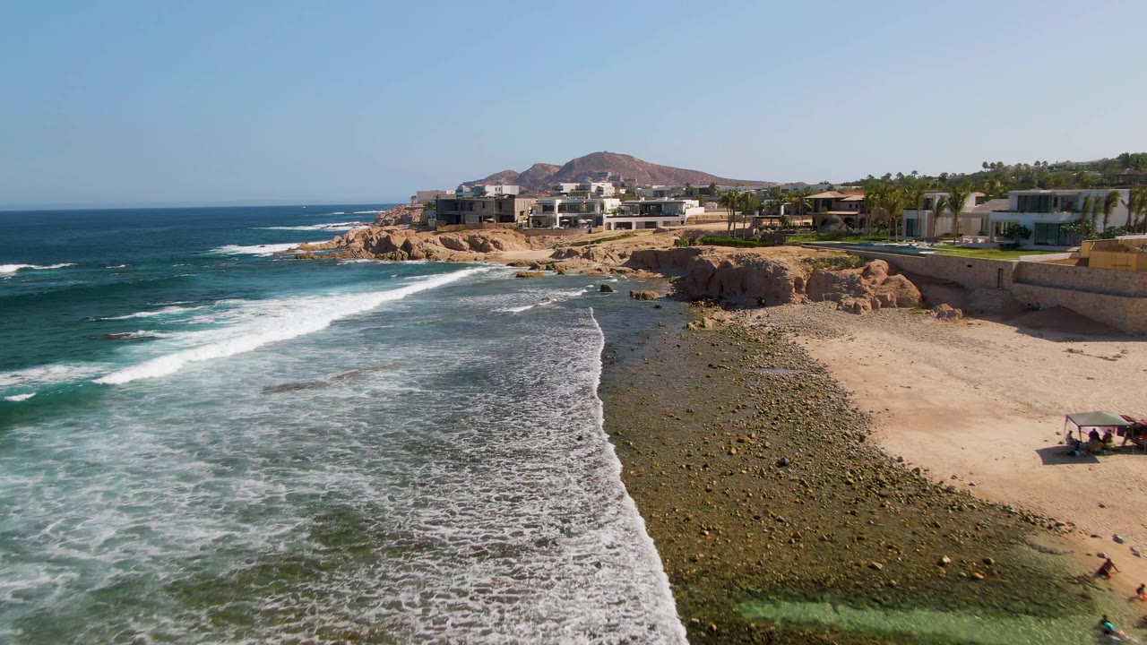 El Tule Beach aerial frame with foamy surf spreading across the reef-like shallows, sand to the right, small beachgoers near the lower right edge, and homes and palms lining the cliff above.