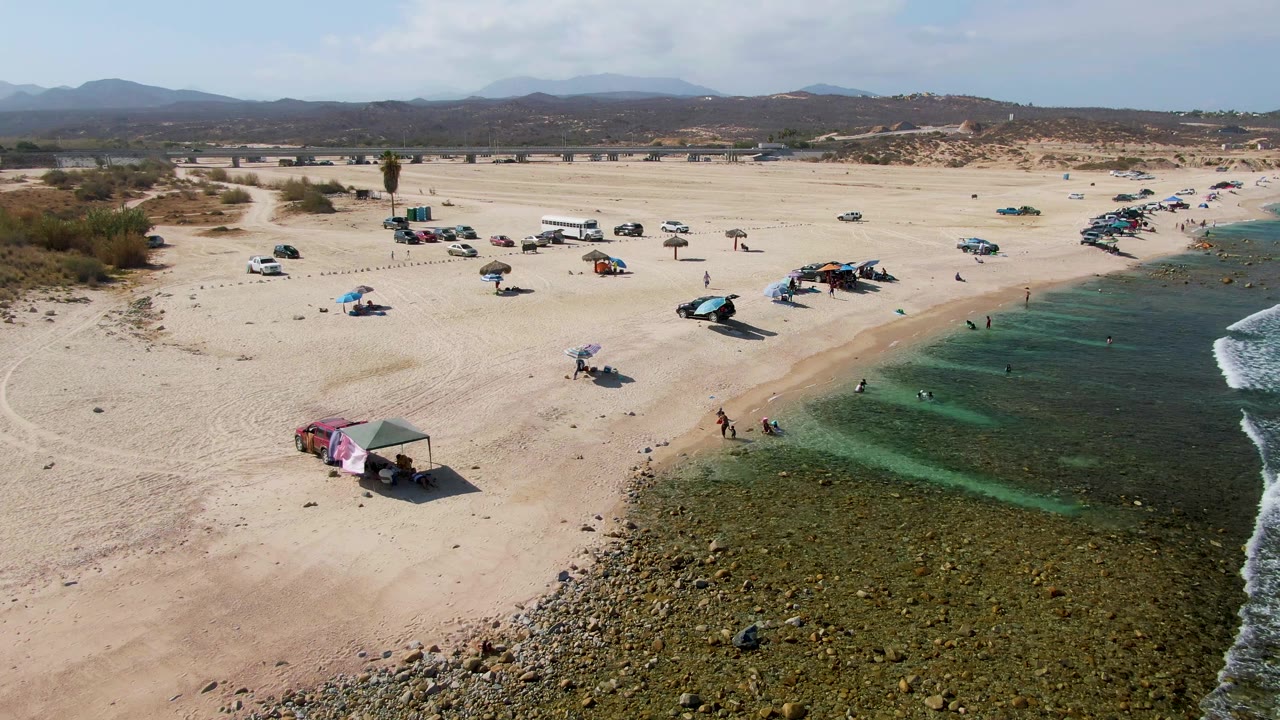 Aerial frame over El Tule Beach Los Cabos shows open sand with parked vehicles and shade tents, people near the shoreline, rocky shallows, and small waves entering from the right.